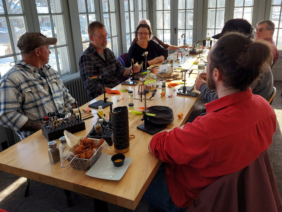 Group of people sitting around a table learning to tie flies with food and drinks in a room with large windows.