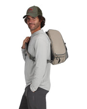 Simms sling pack worn on back, light gray shell with tan side panels