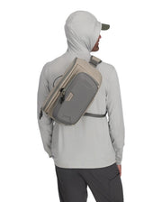 Simms sling pack worn across back and chest, light gray with tan trim