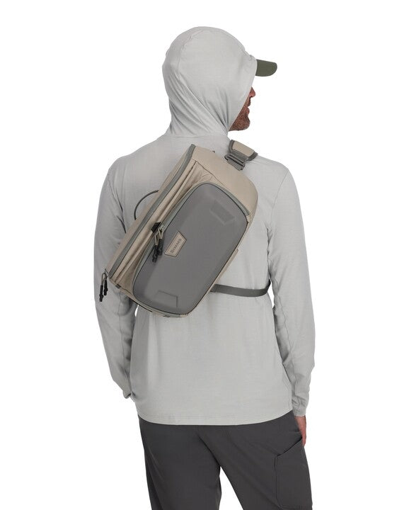 Simms sling pack worn across back and chest, light gray with tan trim