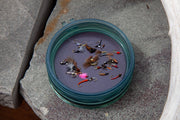 Round fly puck with teal rim, filled with assorted fly fishing lures on a rock