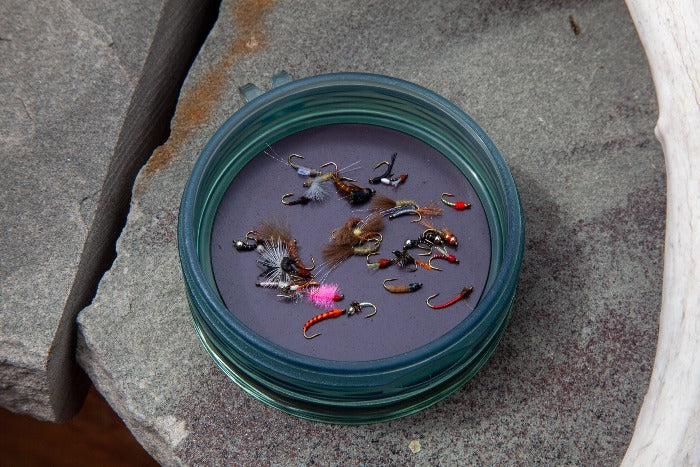 Round fly puck with teal rim, filled with assorted fly fishing lures on a rock