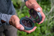 Hands holding a shallow fly puck with teal rim, open container showing fly fishing lures