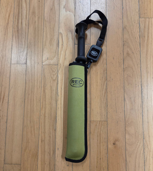 A REC brand wading staff folded and in its olive sheath with a black handle and retractor, lying on a light brown wooden floor. The REC logo is clearly visible on both the sheath and the retractor.