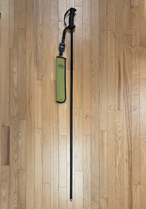 A REC brand wading staff extended and its olive sheath with a black handle and retractor, lying on a light brown wooden floor. The REC logo is clearly visible on both the sheath and the retractor.