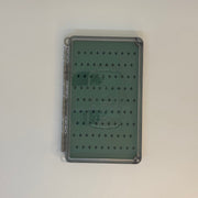 Fly box with closed clear lid and green patterned backing, seen from above