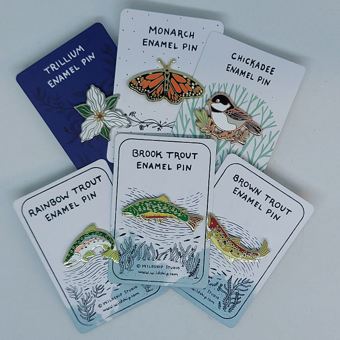 Collection of Wildship Studio enamel pins on cards, featuring trout, chickadee, monarch, trillium.