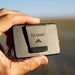 Hand holding a  black 'Fly Caddy' showing the back of the device and its clip with a blurred natural background
