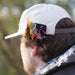 Person wearing a white cap with a fly patch filled with colorful fishing lures clipped on.