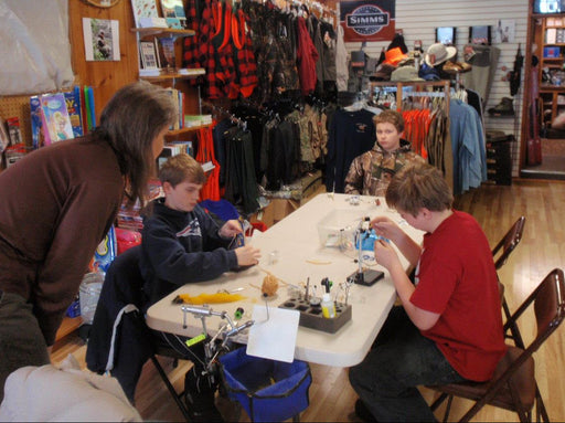 People learning to tie flies at a table in a store with various items and tools around.