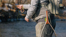 A fly fisherman in a river wearing a light gray waders  and a backpack, holding a fly rod, with a landing net hanging from the waistband.