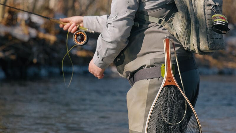 A fly fisherman in a river wearing a light gray waders  and a backpack, holding a fly rod, with a landing net hanging from the waistband.