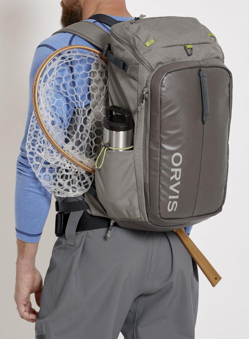 a man wearing the orvis bug out backpack and showing how it can hold a fishing net, water bottle, and more